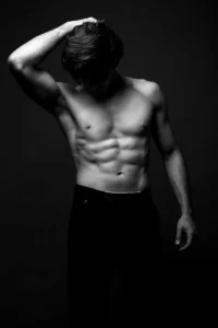 Artistic black and white photograph of a shirtless male model with a muscular build posing in a studio.
