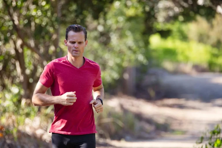 Man in a red shirt jogging on a sunny day along a park pathway surrounded by nature.