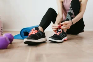 Fit woman tying shoelaces, preparing for indoor workout with yoga mat and dumbbells.