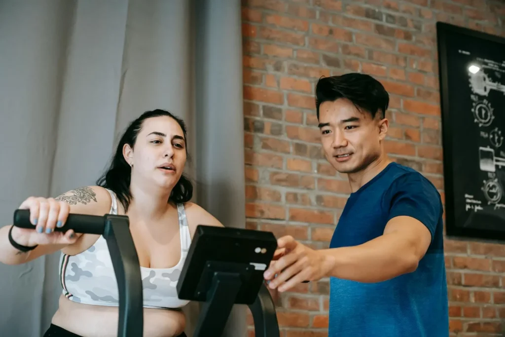Low angle of young ethnic male instructor setting up cycling training apparatus while standing near tired obese woman doing cardio workout in sport club