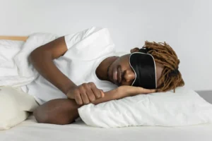 A relaxed African American man sleeping in bed wearing a black sleep mask, promoting restful sleep.