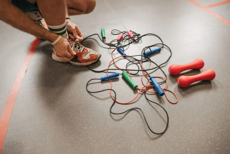 Individual tying shoelaces near colorful jump ropes and red dumbbells on a gym floor.